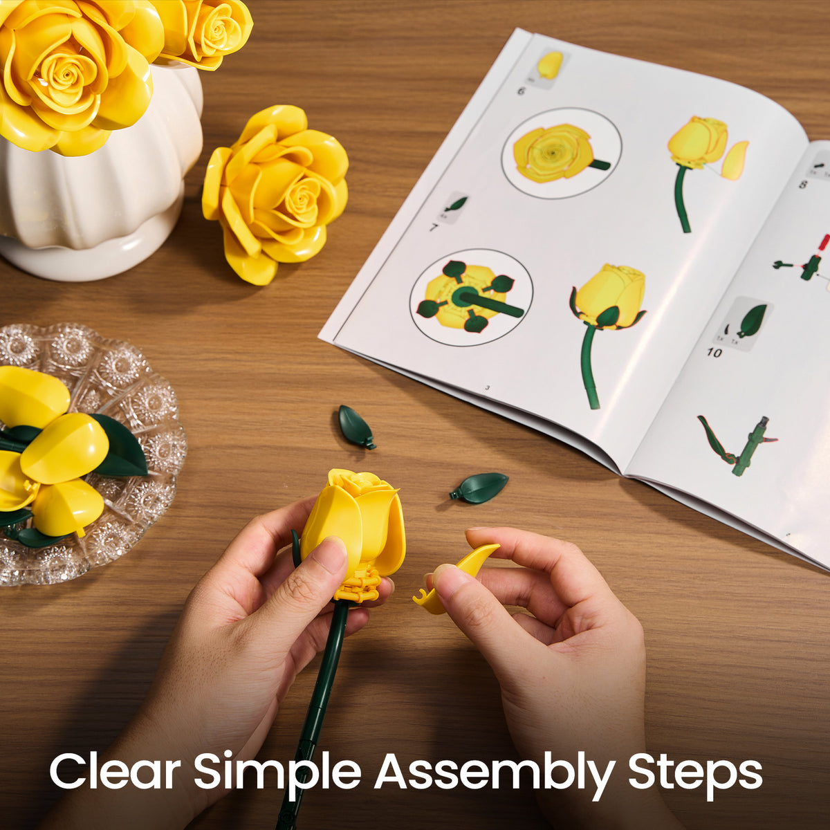 Yellow rose bouquet building set assembly process showing hands attaching flower pieces with instruction guides and loose components on a tabletop.