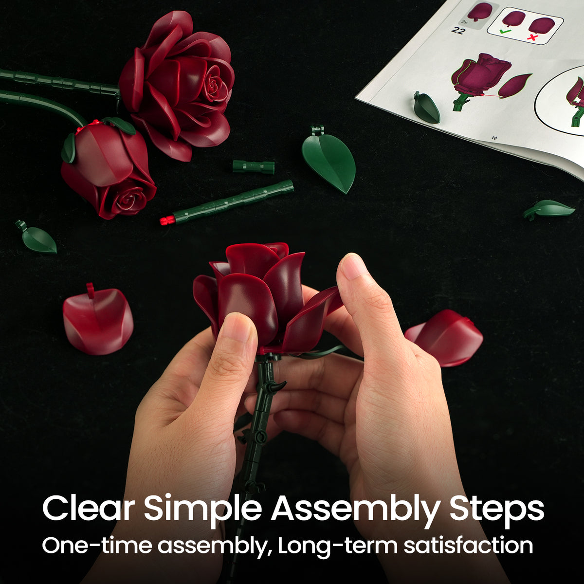 Burgundy rose building bouquet set assembly process showing hands attaching deep red rose petals to a flower stem, with loose rose components, green leaves, and instruction guides displayed on a dark surface.