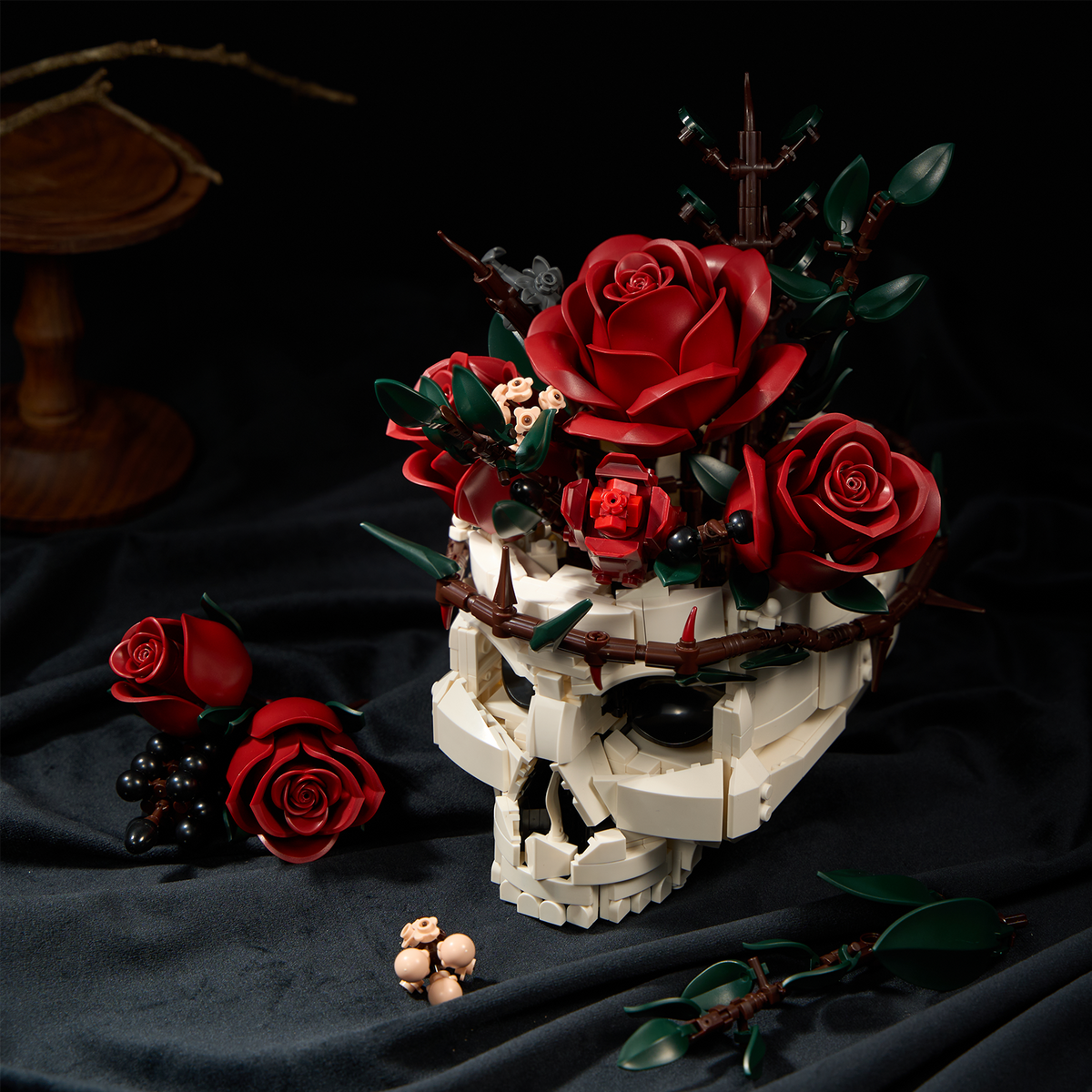 A gothic skull red rose bouquet building block set featuring a white skull base decorated with red roses, green leaves, and dark berry accents, displayed on a black fabric surface with additional rose and floral pieces.