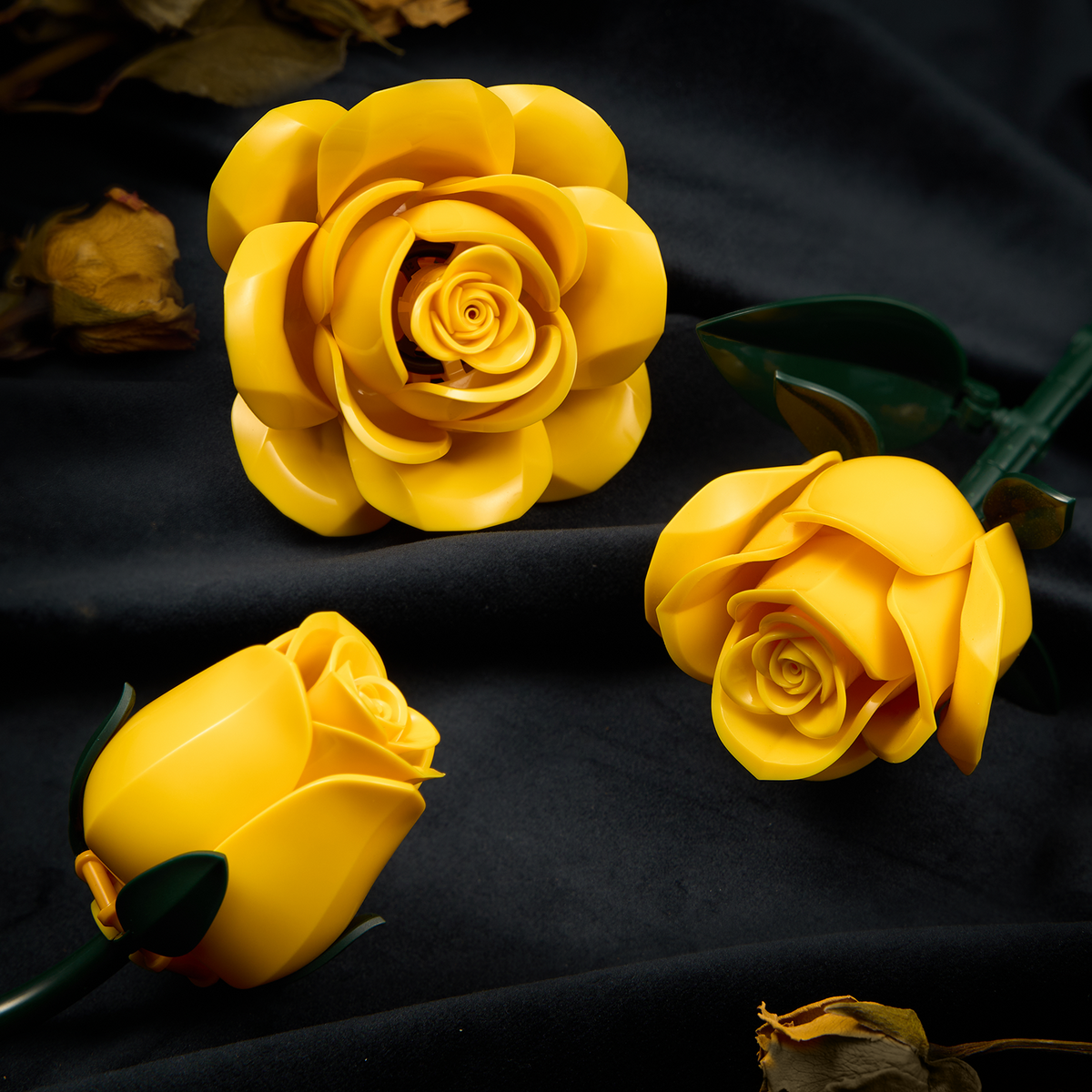 Close-up view of yellow rose building blocks featuring layered petals, realistic rose details, and green leaves arranged on a dark fabric background.