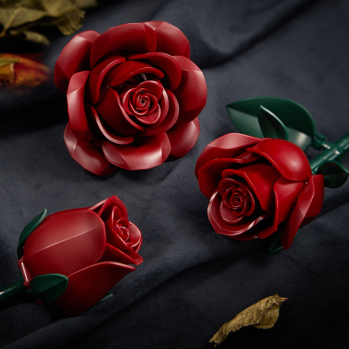 Burgundy rose building bouquet set detail view featuring three deep red rose blooms at different stages, with realistic layered petals, green leaves, and stems arranged on a dark fabric background.
