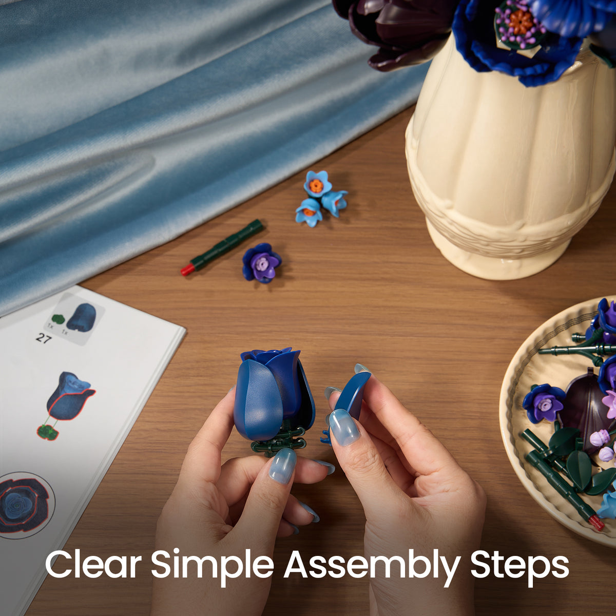 Hands assembling a blue rose from the Blue Flower Bouquet building block set on a wooden table, with instructions and flower pieces nearby