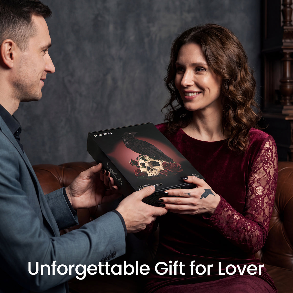 A man and a woman are seated on a brown leather couch, smiling as the man presents a black gift box to the woman. The box features a gothic design with a raven and skull graphic.