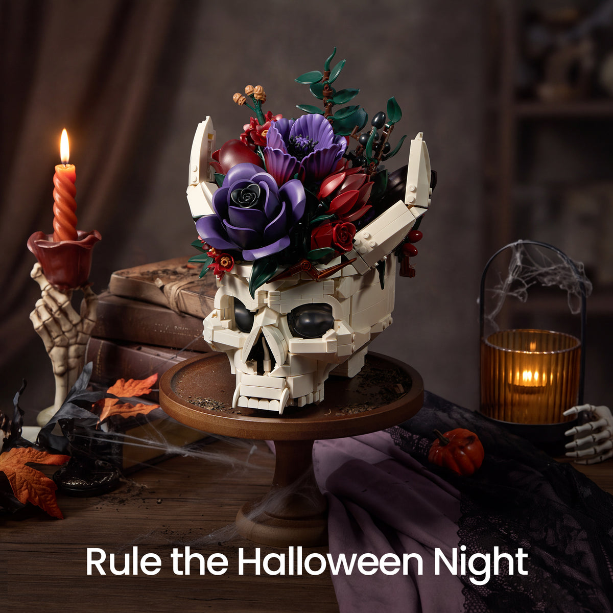 Gothic skull Halloween decor with purple and red flowers displayed beside a lit red candle and small Halloween pumpkins