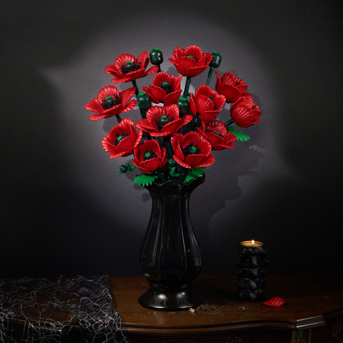 A bouquet of red poppy flowers arranged in a black vase, displayed on a wooden table against a dark textured wall with a candle and decorative elements creating an elegant floral display.