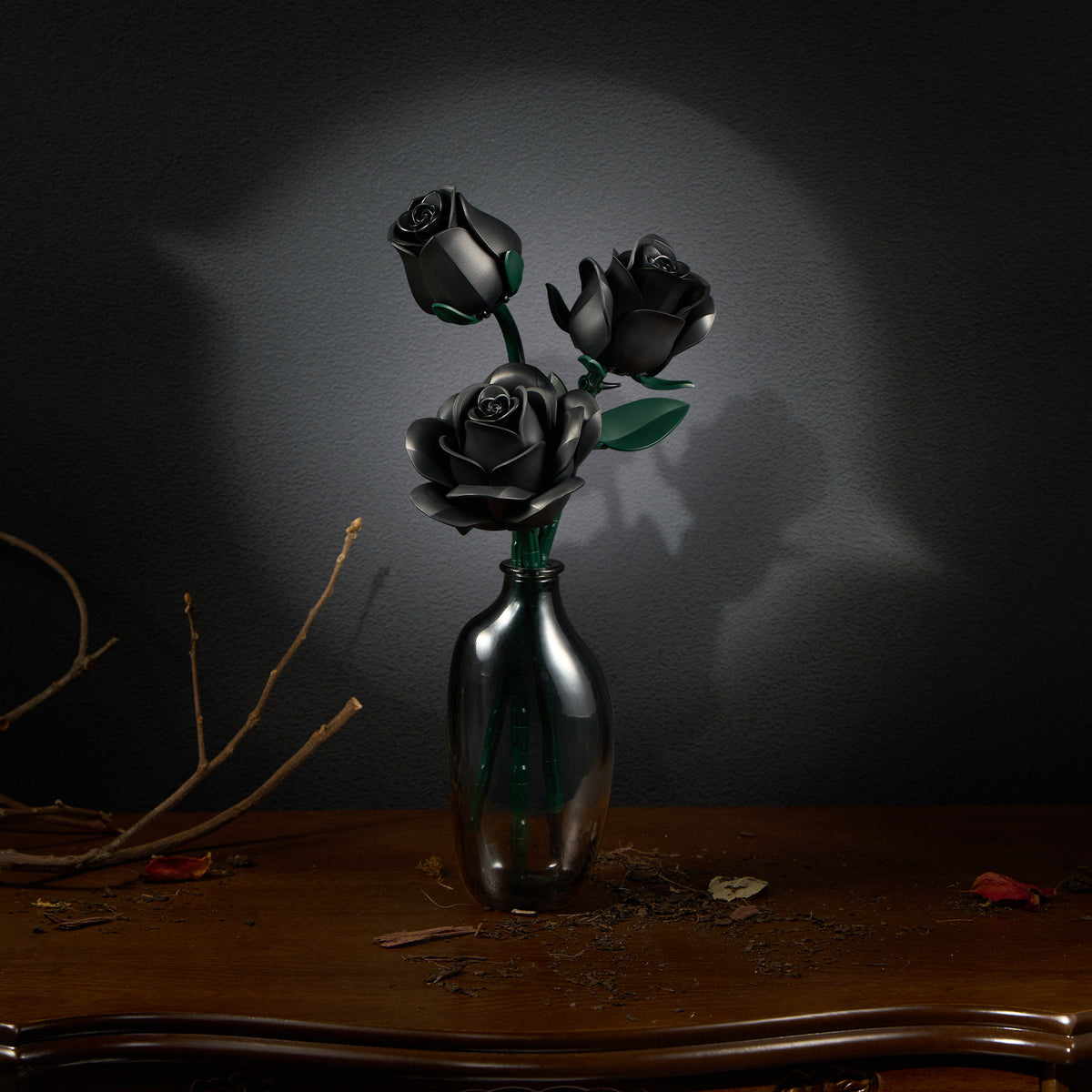 A three black rose building block set arranged in a small metallic vase, displayed on a wooden table against a dark wall, creating a minimalist gothic floral decor scene.